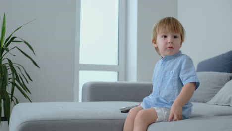 Ein-2-jähriger-Junge-Sitzt-Auf-Einem-Sofa-Und-Schaut-Fern,-Während-Er-Eine-Fernbedienung-In-Der-Hand-Hält.